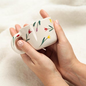 Puede incluir: Una taza de cerámica blanca con un diseño floral en tonos rojos, amarillos y verdes. La taza está siendo sostenida por las manos de una persona.