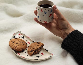 May include: A white ceramic mug with a black, red, and blue pattern. The mug is filled with coffee and is being held by a hand. A white ceramic plate with a black, red, and blue pattern holds two chocolate chip cookies.