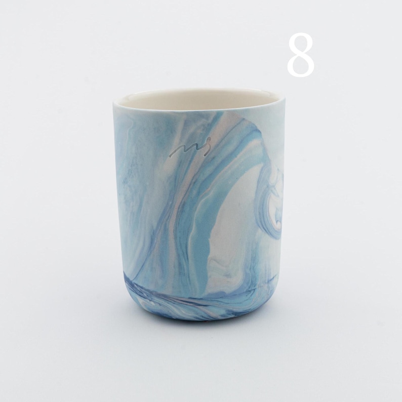 May include: A white ceramic mug with a blue and white marbled design. The mug has a handwritten signature on the side.