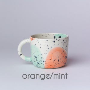 Op de afbeelding: Een witte keramische mok met een gespikkeld glazuur in zwart, oranje en mintgroen. De mok heeft een afgeronde vorm en een handvat.