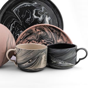Può includere: Una collezione di stoviglie in ceramica con un design marmorizzato. Il set include due tazze, un piatto e un vassoio da portata. La tavolozza dei colori presenta nero, bianco e sfumature di rosa e beige, creando un'estetica moderna.
