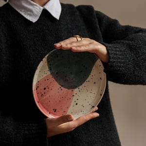 Puede incluir: Un plato de cerámica con fondo blanco y formas abstractas en rosa, verde y negro. El plato tiene un esmalte moteado.
