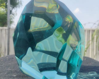Peut inclure: Une sculpture en verre taillé de couleur turquoise avec un design géométrique. La sculpture a une surface à facettes qui reflète la lumière, créant un jeu de couleurs. L'objet est exposé sur une surface sombre.