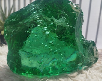 May include: A large, green, glass rock with air bubbles trapped inside. The rock has an irregular shape and is sitting on a white, fluffy surface.