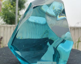 Peut inclure: Une sculpture en verre turquoise facettée de forme géométrique. La sculpture porte une étiquette avec le texte "1525 gr". Le verre est translucide, permettant à la lumière de passer et de créer des reflets.