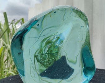 Peut inclure: Une sculpture en verre de forme libre de couleur sarcelle. Le verre d'art transparent a une forme arrondie avec des tourbillons internes de vert et de bleu. La sculpture repose sur une surface sombre, avec un fond de verdure et un mur blanc.