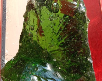 May include: A large, irregularly shaped piece of green glass with air bubbles trapped inside. The glass has a smooth, shiny surface and is translucent.