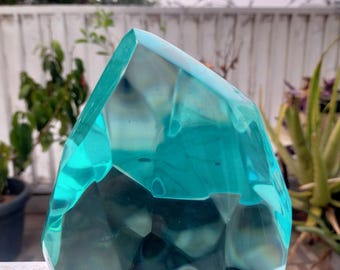 Peut inclure: Une sculpture en verre facetté de couleur turquoise, au design géométrique. Cette œuvre d'art transparente a une apparence cristalline, captant la lumière et reflétant l'environnement. La sculpture est exposée à l'extérieur.