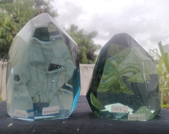 Peut inclure: Deux gros presse-papiers en verre transparent facetté avec une teinte verdâtre. Les presse-papiers sont en forme de cristal et ont une surface texturée. Les presse-papiers sont posés sur une surface noire. Le plus grand presse-papiers porte l'inscription "10x4.5x14" sur le fond. Le plus petit presse-papiers porte l'inscription "7x4x8" sur le fond.