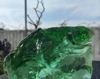 May include: A large, irregularly shaped chunk of translucent green glass. The glass has a rough, textured surface with many facets, catching the light and creating a sparkling effect. The background shows a blurred sky and a fence.