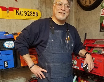May include: A man wearing a blue denim apron with multiple pockets and a pair of jeans stands in a workshop. He is holding a small black device in his right hand. The workshop has various tools and equipment, including a red toolbox with the text "LiPo Battery Storage" and a silver toolbox with the text "IX 12 SPEKTRUM".