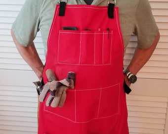 May include: A red canvas apron with multiple pockets and white stitching. The apron has black straps and is worn by a person with a beard.