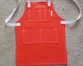 May include: Orange canvas apron with white straps and white stitching. The apron has two large pockets and a smaller pocket on the chest. The name "Owyn" is embroidered on the chest pocket.
