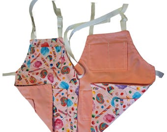 May include: Two aprons. One apron features a pink candy pattern with lollipops, cotton candy, and candy canes. The other apron is solid peach. Both aprons have white straps and pockets.