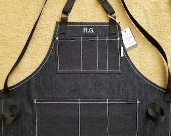 May include: A dark blue denim apron with white stitching and multiple pockets. The apron has adjustable straps and a personalized embroidered name "R.G."