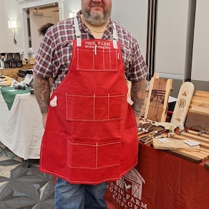 May include: A man wearing a red apron with the text "Chik Farm Woodworking" is standing in front of a table displaying wooden crafts. The table has a red tablecloth with the same logo printed on it.