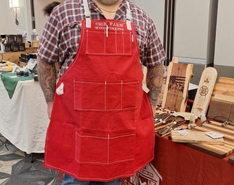 May include: A man wearing a red apron with the text "Chik Farm Woodworking" is standing in front of a table displaying wooden crafts. The table has a red tablecloth with the same logo printed on it.