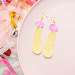 May include: A pair of dangle earrings with a pink top and a yellow, patterned bottom. The earrings are on a pink plate, with colorful streamers and disco ball decorations in the background. The earrings have gold-colored hooks.