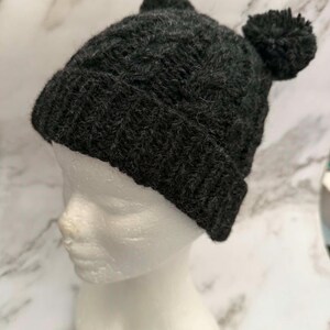 May include: A dark charcoal grey knit beanie featuring a ribbed cuff and two matching pom-poms on the top. The hat is crafted with a textured cable knit pattern throughout the main body and is displayed on a white mannequin head.