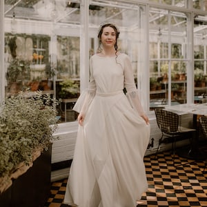 Puede incluir: Una novia con un vestido de novia blanco con falda larga y mangas acampanadas. Está de pie en un invernadero con un suelo a cuadros y un techo blanco.