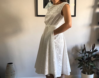 Puede incluir: Vestido sin mangas de color blanco roto con un diseño texturizado. El vestido presenta un corpiño ajustado, una cintura definida con cinturón y una falda acampanada. Tiene bolsillos y llega hasta la pantorrilla. El vestido está hecho de una tela suave y aterciopelada.