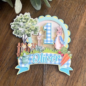 Op de afbeelding: Een blauwe en witte geruite taarttopper met een konijn, een boom, een hek, een gieter, wortels en het nummer 1. De topper heeft ook een blauwe banner met de naam "Oliver" erop.
