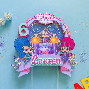 Może przedstawiać: Topper na tort w kolorze fioletowym i niebieskim z dwoma postaciami z kreskówki z serialu 'Shimmer and Shine'. Postacie stoją przed fioletowym i złotym pałacem. Topper zawiera również baner z napisem 'Happy Birthday' i baner z napisem 'Lauren'.