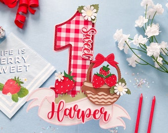 May include: A red and white gingham number one cake topper with a basket of strawberries, a white flower, and the word "Sweet" written on it. The topper also includes a pink banner with the name "Harper" written on it.