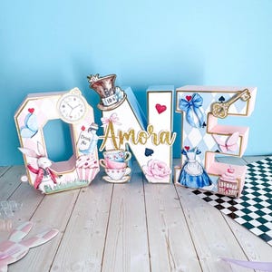 Op de afbeelding: Een decoratief "ONE" bord met een Alice in Wonderland thema. De letters zijn roze, blauw en goud, met illustraties van een konijn, theekopjes, een klok en een jurk. De naam "Amora" is geschreven in gouden glitter.
