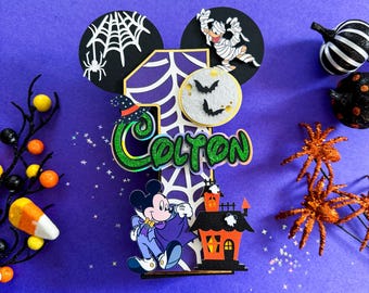 May include: A purple and black Halloween-themed cake topper with a number one, Mickey Mouse dressed as a vampire, a mummy, a spider web, and the name Colton.