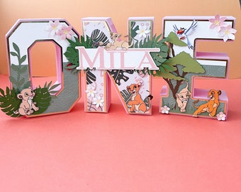 Può includere: Lettere in cartone rosa e verde che compongono la parola "ONE" con uno striscione rosa che dice "MILA". Le lettere sono decorate con immagini di leoni e fogliame della giungla.