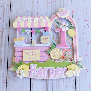 May include: A pink and yellow lemonade stand cake topper with a pink number one, a sign that says "Lemonade 25¢", and the name "Harper" in pink letters.