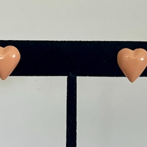 May include: A pair of peach-colored heart-shaped stud earrings. The earrings are displayed on a black velvet earring holder against a white background. The earrings are a solid color and have a glossy finish.