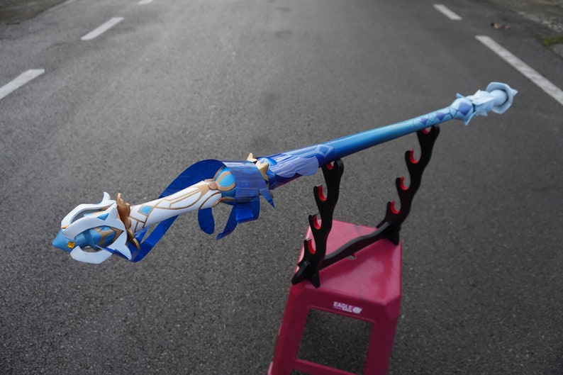 Peut inclure: Une &eacute;p&eacute;e bleue et blanche avec des accents dor&eacute;s est expos&eacute;e sur un support d'&eacute;p&eacute;e noir. L'&eacute;p&eacute;e repose sur un tabouret en plastique rose.