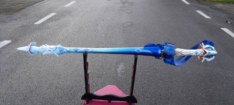 Peut inclure: Une &eacute;p&eacute;e bleue et blanche avec une poign&eacute;e dor&eacute;e et un ruban bleu. L'&eacute;p&eacute;e est sur un support noir.