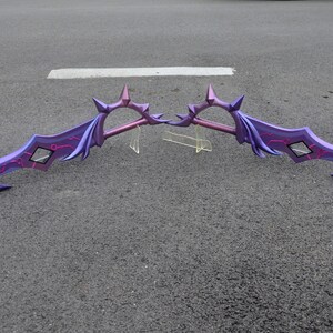 May include: A pair of purple and pink fantasy swords with intricate details and a spiked design. The swords are laid out on a gray asphalt surface.