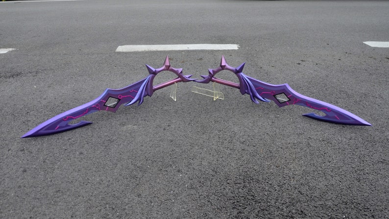 May include: A pair of purple and pink fantasy swords with intricate details and a spiked design. The swords are laid out on a gray asphalt surface.