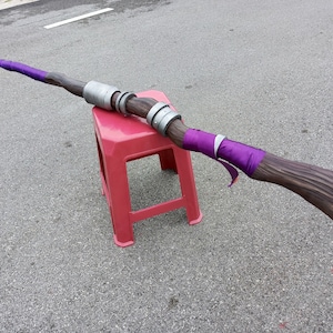 May include: A long staff featuring a brown wood-textured finish, accented with vibrant purple fabric wraps at both ends. The center of the staff is adorned with two metallic silver cylindrical rings, creating a distinct contrast against the dark wood.