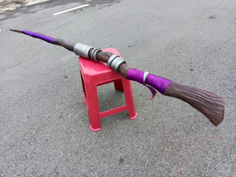 May include: A long staff featuring a brown wood-textured finish, accented with vibrant purple fabric wraps at both ends. The center of the staff is adorned with two metallic silver cylindrical rings, creating a distinct contrast against the dark wood.