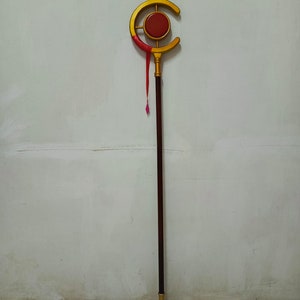 May include: A long staff featuring a dark brown cylindrical shaft with a gold-colored base and a top ornament shaped like a crescent moon. The crescent surrounds a central red circular disc, and a thin red ribbon hangs from the gold frame.