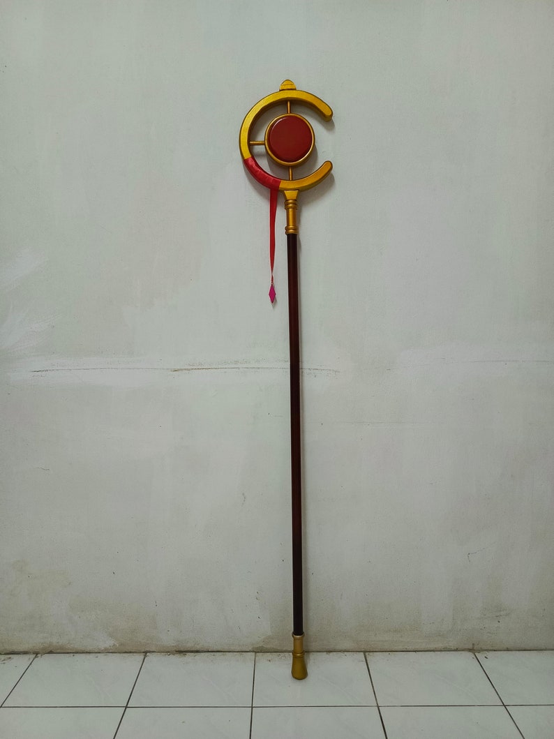 May include: A long staff featuring a dark brown cylindrical shaft with a gold-colored base and a top ornament shaped like a crescent moon. The crescent surrounds a central red circular disc, and a thin red ribbon hangs from the gold frame.