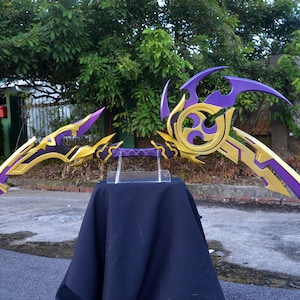 May include: A pair of decorative fantasy-style bows with gold and purple paint. The bows are displayed on a black cloth stand.