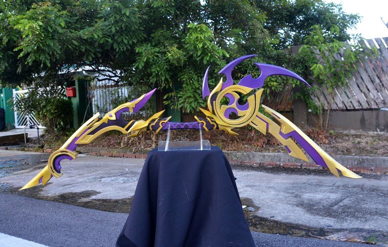 May include: A pair of decorative fantasy-style bows with gold and purple paint. The bows are displayed on a black cloth stand.