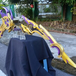May include: A purple and gold bow and arrow prop with intricate details. The bow is curved and has a sharp point. The arrow is straight and has a feathered end.