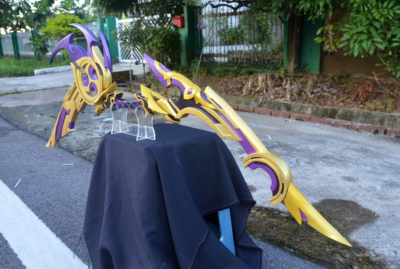 May include: A purple and gold bow and arrow prop with intricate details. The bow is curved and has a sharp point. The arrow is straight and has a feathered end.