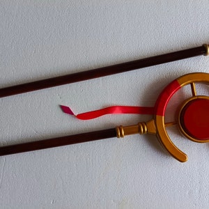 May include: A two-piece staff featuring a dark brown shaft with gold-colored accents. The top section includes a circular gold frame surrounding a solid red center disc, with a red ribbon attached to the side and a small gold finial at the end.