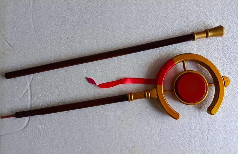 May include: A two-piece staff featuring a dark brown shaft with gold-colored accents. The top section includes a circular gold frame surrounding a solid red center disc, with a red ribbon attached to the side and a small gold finial at the end.