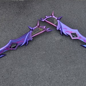 May include: Two purple and pink fantasy swords with intricate designs and sharp points. The swords are crossed and lying on a gray surface.