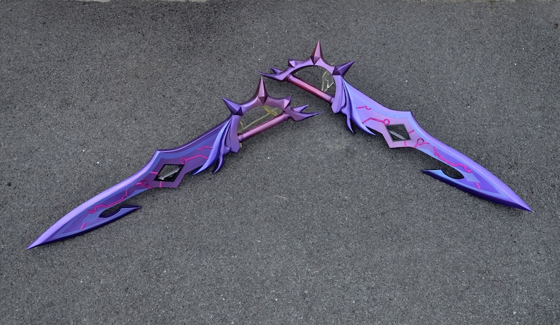 May include: Two purple and pink fantasy swords with intricate designs and sharp points. The swords are crossed and lying on a gray surface.