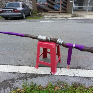 May include: A long, staff-like prop featuring a textured brown wood-grain finish, accented with metallic silver bands and vibrant purple fabric wraps. A long purple fabric strip hangs from the center, ending in a small, pointed red charm.